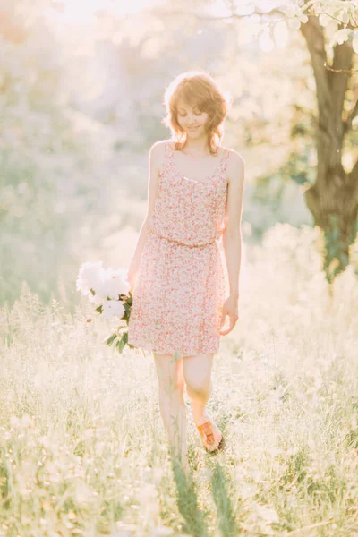 Vacker ung vit blond kvinna i lätt klänning med bukett av vita pioner, promenader i sommarfält eller trädgård i solnedgången. Kvinna med blommor utomhus — Stockfoto