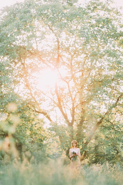 Belle jeune femme blonde caucasienne en robe légère tenant un bouquet de pivoines blanches, se promenant dans un champ d'été ou un jardin au coucher du soleil. Femme avec des fleurs à l'extérieur — Photo