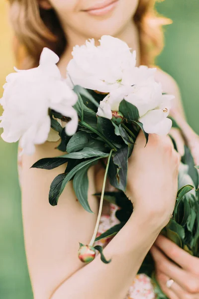 Mulher loira branca bonita em vestido leve segurando buquê de peônias brancas, andando no campo de verão ou jardim ao pôr do sol. Mulher com flores ao ar livre — Fotografia de Stock