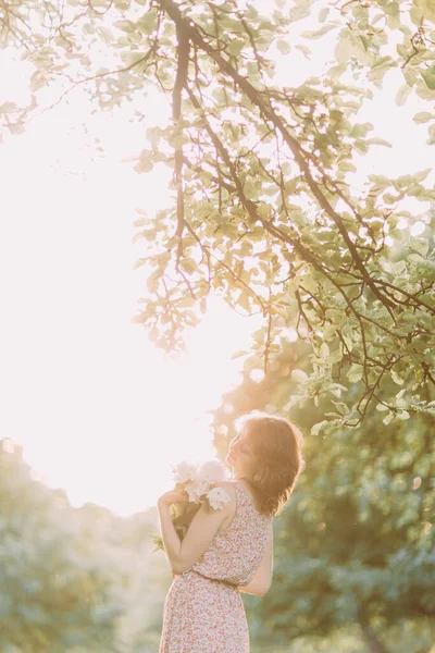 白い牡丹の花束を保持光のドレスで美しい若い白人ブロンドの女性は、夏のフィールドや日没の庭を歩く。屋外で花を持つ女性 — ストック写真