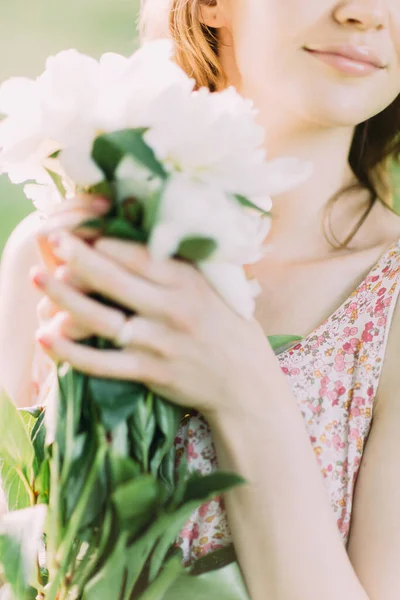 Mulher loira branca bonita em vestido leve segurando buquê de peônias brancas, andando no campo de verão ou jardim ao pôr do sol. Mulher com flores ao ar livre — Fotografia de Stock