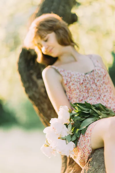 Mulher loira branca bonita em vestido leve segurando buquê de peônias brancas, andando no campo de verão ou jardim ao pôr do sol. Mulher com flores ao ar livre — Fotografia de Stock