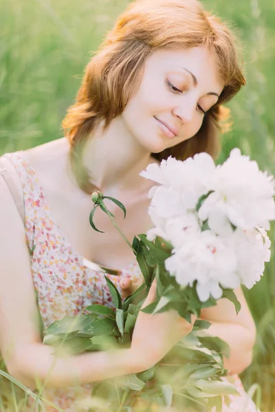 Beautiful young Caucasian blond woman in light dress holding bouquet of white peonies, walking in summer field or garden in sunset. Woman with flowers outdoors — Stock Photo, Image