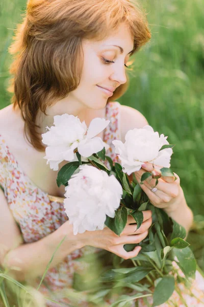 Mulher loira branca bonita em vestido leve segurando buquê de peônias brancas, andando no campo de verão ou jardim ao pôr do sol. Mulher com flores ao ar livre — Fotografia de Stock