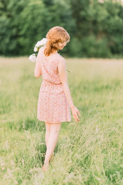 Mulher loira branca bonita em vestido leve segurando buquê de peônias brancas, andando no campo de verão ou jardim ao pôr do sol. Mulher com flores ao ar livre — Fotografia de Stock