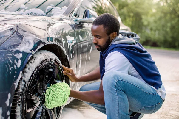 Mladý pohledný Afričan se zelenou houbou, myje auto pěnou. Čištění moderních ráfků luxusního modrého elektromobilu v samoobsluze mytí aut venku. — Stock fotografie