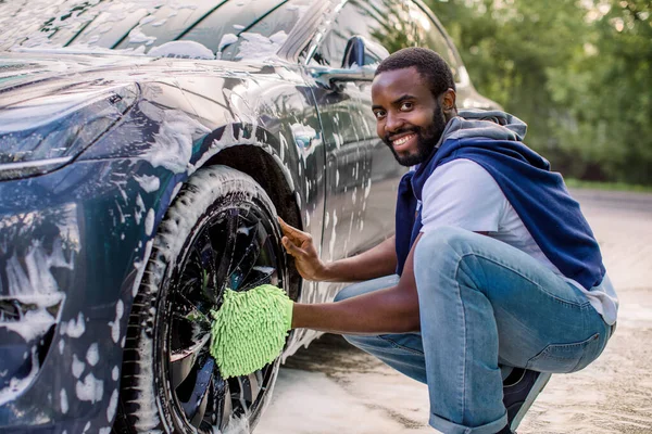 Widok z boku przystojnego młodego Afrykanina używającego zielonej rękawicy z mikrofibry do czyszczenia obręczy jego nowoczesnego samochodu na zewnątrz. Luksusowy niebieski samochód elektryczny w piance mydlanej na zewnątrz w myjni samochodowej. — Zdjęcie stockowe