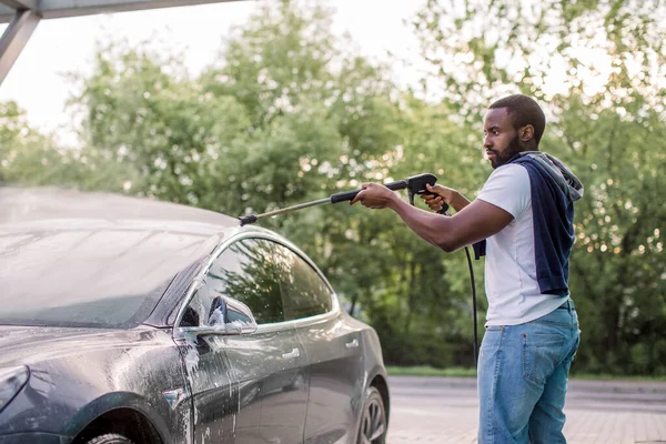 Africano joven enfocado en el hombre en ropa casual lavando su coche eléctrico moderno con una pistola de agua en la estación de auto-servicio de lavado de coches al aire libre, enjuagar la espuma —  Fotos de Stock