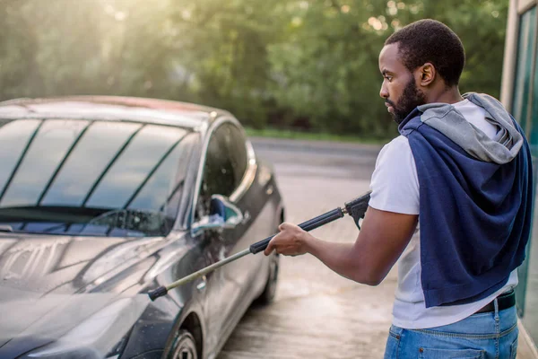 Araba yıkama servisi. Beyaz tişörtlü ve kot pantolonlu Afrikalı adam, açık hava oto yıkamada su tabancasıyla mavi elektrikli araba yıkıyor.. — Stok fotoğraf