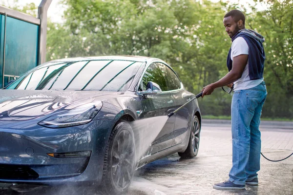 Snygg afrikansk kille i t-shirt och jeans tvättar sin blå moderna elektriska lyxbil, sköljer tvålen med högtrycksvatten jet. Manuell biltvätt med trycksatt vatten i biltvätt utanför. — Stockfoto