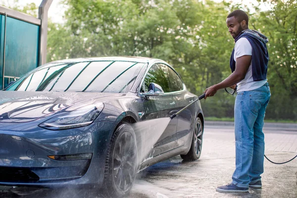 Bello ragazzo africano in t-shirt e jeans che lava la sua moderna auto di lusso elettrica blu, risciacquando il sapone con getto d'acqua ad alta pressione. Autolavaggio manuale con acqua pressurizzata in autolavaggio esterno. — Foto Stock