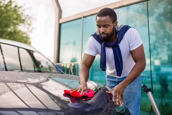 Afro-Amerikan bir adamın lüks elektrikli arabasının kaputunu temizlerken çekilmiş yatay görüntüsü. Kırmızı mikrofiber bezle, oto yıkamada. — Stok fotoğraf