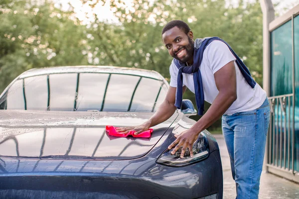 Genç mutlu gülümseyen Afrikalı adam gelecekteki elektrikli araba kaputunu ve farlarını kırmızı mikrofiber bezle temizliyor, kameraya bakıyor. Açık hava oto yıkama servisi.. — Stok fotoğraf