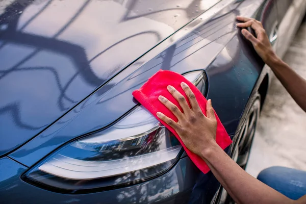 Closeup recortou a imagem das mãos do jovem homem africano, lavando e limpando seu carro azul moderno com pano de microfibra vermelha, limpando capuz e faróis. Lavagem de carro conceito ao ar livre . — Fotografia de Stock