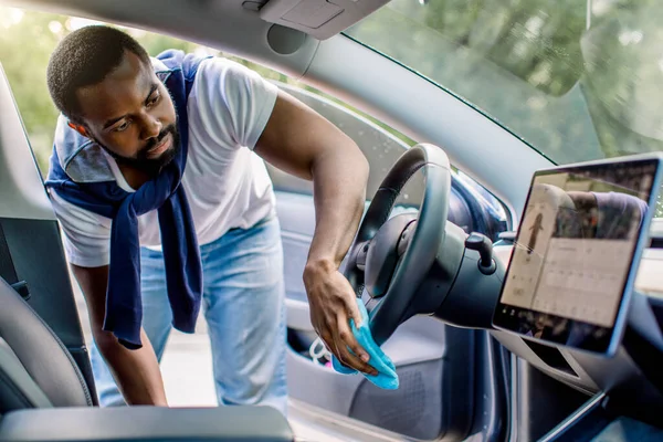 Pulizia di interni auto, auto concetto di dettaglio. Giovane uomo africano in abbigliamento casual lava un interno auto, volante auto con vestiti in microfibra blu, in un self-service di lavaggio auto all'aperto — Foto Stock