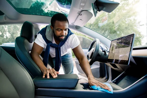 Cleaning service, car interior washing. Handsome African man in casual clothes, washes a car interior with microfiber cloth in his modern electric car with self-steering system