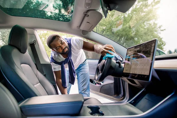 Servicio de limpieza, concepto de detalle del coche. Joven hombre de piel oscura en camiseta y jeans, lavando el interior de un coche, volante de coche con ropa de microfibra, en su moderno coche futurista de alta tecnología — Foto de Stock