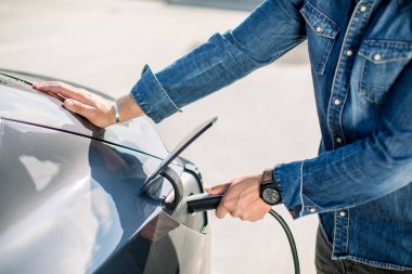 Kotlu tişörtlü bir adamın ve şarj istasyonundaki elektrikli arabasının elektrik kablosu fişe takılı olduğu kesikli görüntüyü kapat. Elektrikli arabalar, gelecekteki araba konsepti