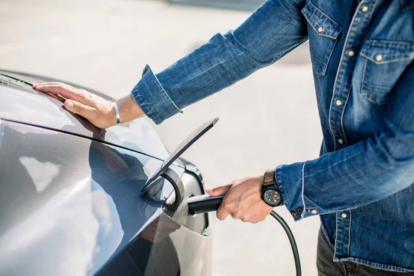 Kotlu tişörtlü bir adamın ve şarj istasyonundaki elektrikli arabasının elektrik kablosu fişe takılı olduğu kesikli görüntüyü kapat. Elektrikli arabalar, gelecekteki araba konsepti — Stok fotoğraf