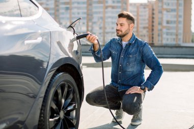 Yakışıklı sakallı adam yeni modern elektrikli arabasının yanında oturuyor ve şarj aletinin fişini çekiyor, araba şarj istasyonunda şarj olurken