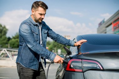 Kot pantolonlu, sakallı genç bir adamın yan görüntüsü. Lüks modern elektrikli arabasını şarj etmek için araba prizine tel takıyor. Şehirdeki şarj istasyonunda.