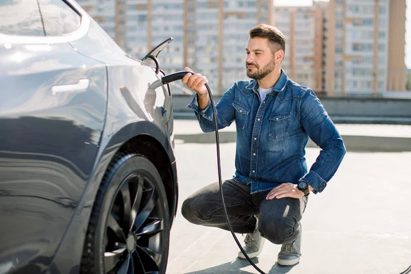 Hansome barbudo tipo sentado cerca de su nuevo coche eléctrico moderno y la celebración de enchufe del cargador, mientras que el coche se está cargando en la estación de carga —  Fotos de Stock