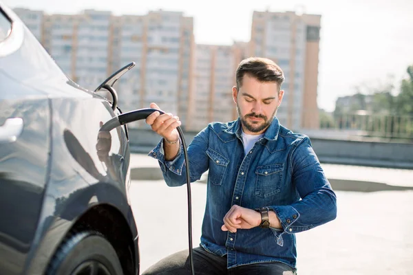 Homem barbudo Hansome sentado perto de seu novo carro elétrico moderno e segurando o plugue do carregador, enquanto o carro está carregando na estação de carregamento. Um homem olha para o seu relógio de pulso — Fotografia de Stock