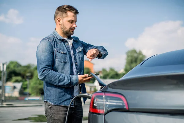Elektrikli arabanın yanında duran beyaz sakallı genç adam saate ve akıllı telefona bakıyor. Elektrikli arabası elektrikli araçlar için şarj istasyonunda çalışıyor.. — Stok fotoğraf