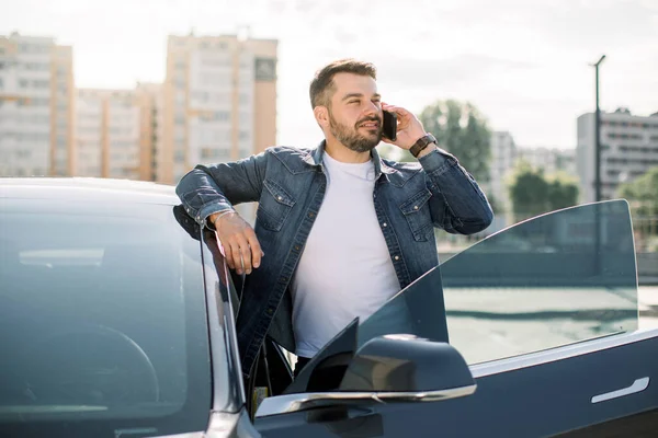 Beau jeune homme barbu caucasien parlant sur le téléphone portable et souriant tout en s'appuyant sur sa nouvelle voiture de luxe moderne, debout à l'extérieur sur le fond de nouveaux bâtiments — Photo