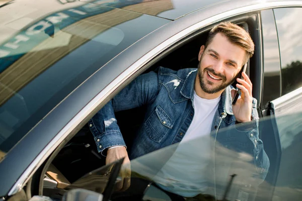 Guapo hombre de barba caucásica con camisa vaquera casual, sentado en su nuevo coche moderno, hablando por teléfono celular, sonriendo y mirando a la cámara. El dueño del hombre feliz está probando su coche nuevo — Foto de Stock