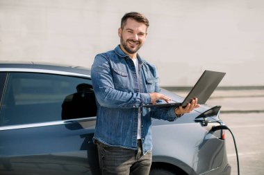 Genç yakışıklı adam dizüstü bilgisayarda çalışıyor, bekliyor, lüks elektrikli arabası şarj olurken, ücretini öderken, elektrik kablosu takılı bir şekilde şehir merkezinin yakınında dururken.