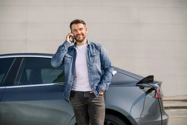 Smiling Caucasian man in casual wear talking phone while leaning on his luxury new electric car, standing on the charging station for charging a car. Konsep pengisian mobil listrik — Stok Foto