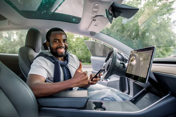 Pembalap pria Afrika tersenyum yang ceria duduk di mobil listrik berteknologi tinggi barunya dengan sistem avtopilot self-driving yang ditingkatkan, memegang smartphone-nya dan menunjukkan jempol ke atas — Stok Foto