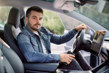 Otomatik sürüş sistemi olan yeni nesil elektro aracın test sürüşü. Yakışıklı, sakallı beyaz adam yeni modern arabanın direksiyonunda oturuyor ve kameraya bakıyor. Yenilik Teknolojisi