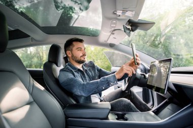 Yakışıklı beyaz adam modern otomatik direksiyonlu elektrikli araçta oturuyor, akıllı telefonunda mobil uygulama kullanıyor, park halinde, şarj oluyor.