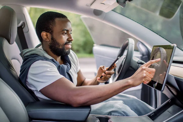 Young excited African man working with car touchscreen display and mobile car app, while sitting in his modern electric futuristic vehicle with self-steering system