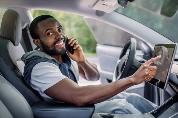 아프리카계 미국인젊은 남성이 편안 한 현대식 전기 자동차의 선실에 앉아 전화를 하고 있는 모습과 자기가 운전하는 자동 운전 스크린에 손을 대고 있는 모습. 새 차를 시험하는 모습 — 스톡 사진