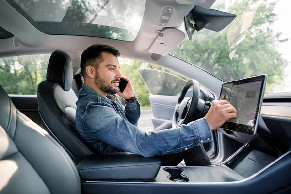 Testing a new electric futuristic car with self driving system. Side view of satisfied Caucasian man in casual jeans shirt sitting in modern car, talking phone and toushing navigation autopilot screen