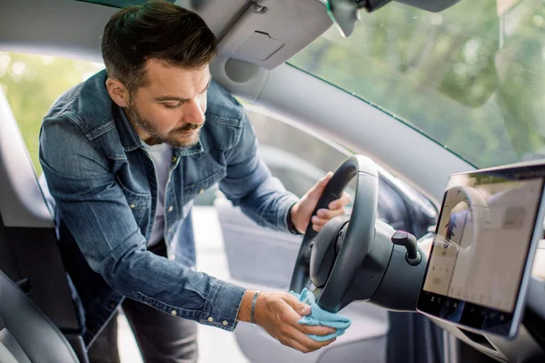 Cleaning of car interior, car detailing concept. Young Caucasian man in casual wear washes a car interior, car steering wheel with blue microfiber clothes, in a outdoor car wash self service