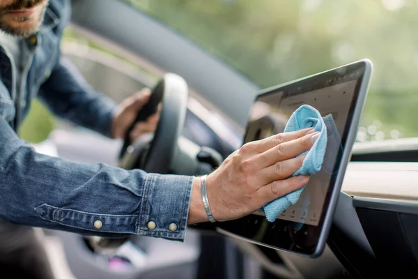 Primer plano plano recortado de la mano de un joven con camisa vaquera, limpiando el polvo de la pantalla táctil con tela de microfibra, en el interior del coche eléctrico de dirección automática moderna. Céntrate en la mano —  Fotos de Stock