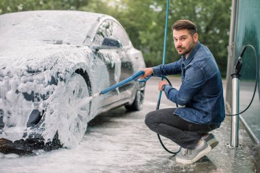 Araba yıkama konsepti. Köpüklü modern lüks elektrikli araba. Yüksek basınçlı jeti olan genç beyaz bir adam araba yıkama istasyonunda sabun püskürtüyor.