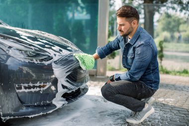 Araba yıkama servisinde araba yıkama servisi. Yakışıklı sakallı adam, günlük giysiler içinde, araba radyatörü ızgarası ve yeni elektrikli arabasının kaputunu köpükle ve süngerle yıkıyor..