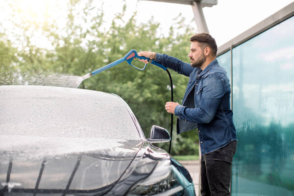 Young handsome Caucasian man cleaning his luxury car with foam spray outside at self car wash station. Casual bearded guy washing his car with high pressure sprayer and foam.
