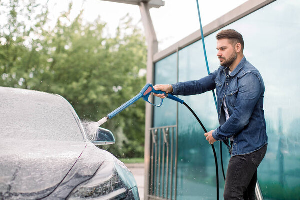 Handsome young man in casual wear is spraying cleaning foam to a modern car in outdoor self car wash service with a high pressure washer. Modern car and washing outdoors