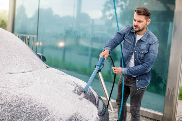 Yüksek basınçlı jet kullanarak arabayı sabun köpüğüyle temizleyin. Yakışıklı sakallı genç adam yüksek basınç altında arabasını dışarıda köpükle yıkıyor.. — Stok fotoğraf