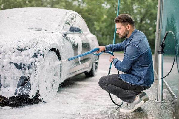 Araba yıkama konsepti. Köpüklü modern lüks elektrikli araba. Yüksek basınçlı jeti olan genç beyaz bir adam araba yıkama istasyonunda sabun püskürtüyor. — Stok fotoğraf
