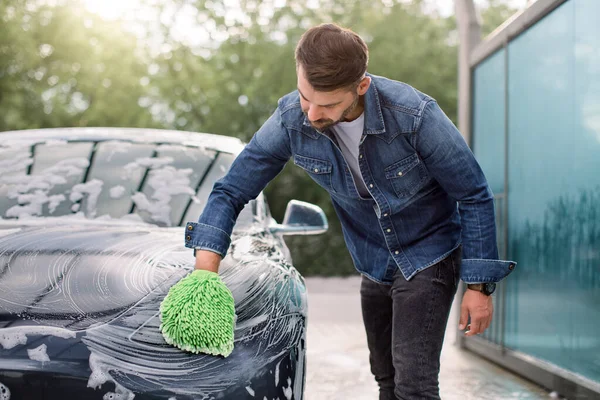 Az autótisztítás horizontális kilátása a szabadban. Jóképű fiatal fehér férfi alkalmi viseletben mossa modern elektromos autóját zöld kesztyű szivaccsal és habbal. — Stock Fotó
