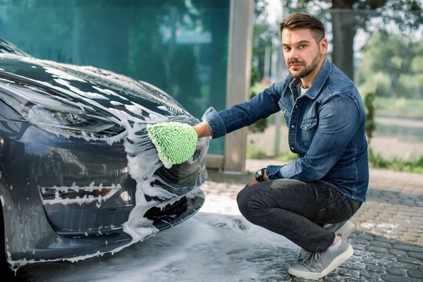 Närbild foto av ung vit man rengöring huva av lyx elbil med skum och grön pensel vante. Biltvätt och detaljer. Biltvätt koncept. Biltvätt på självtvättstationen utomhus — Stockfoto