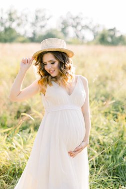Gülümseyen, hamile bir kadın, beyaz elbise ve hasır şapka giyiyor, yaz tarlasında dinleniyor. Açık havada poz vermek. Annelik. Beyaz elbiseli hamile güzel bir kadının açık hava portresi