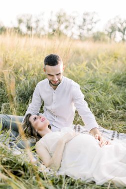 Yakışıklı, sakallı, beyaz bir adam sıcak yaz akşamında çimlerin üzerinde dinlenen büyüleyici hamile karısına bakıyor.